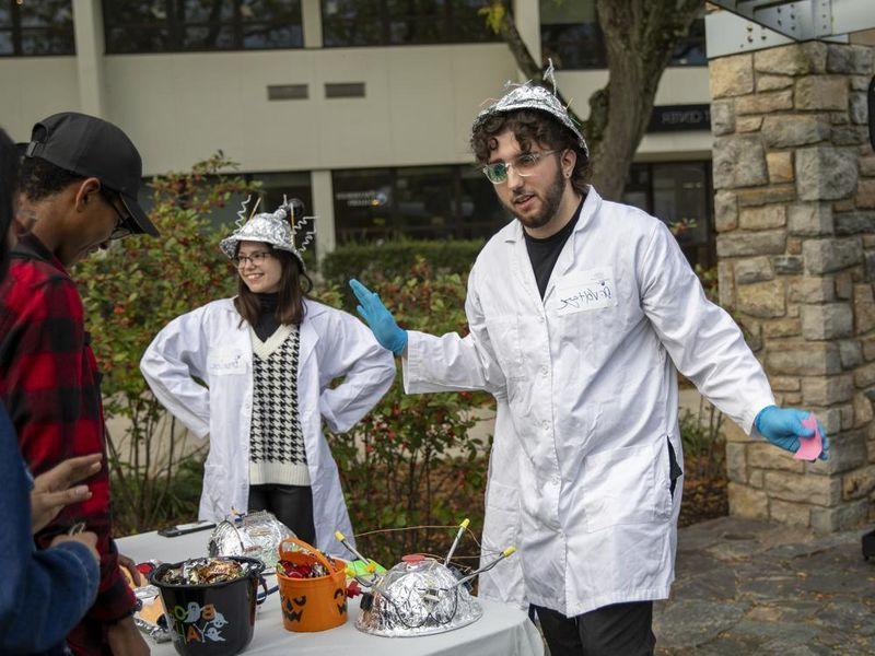 Students wearing lab coats and tinfoil hats conduct experiments with onlookers